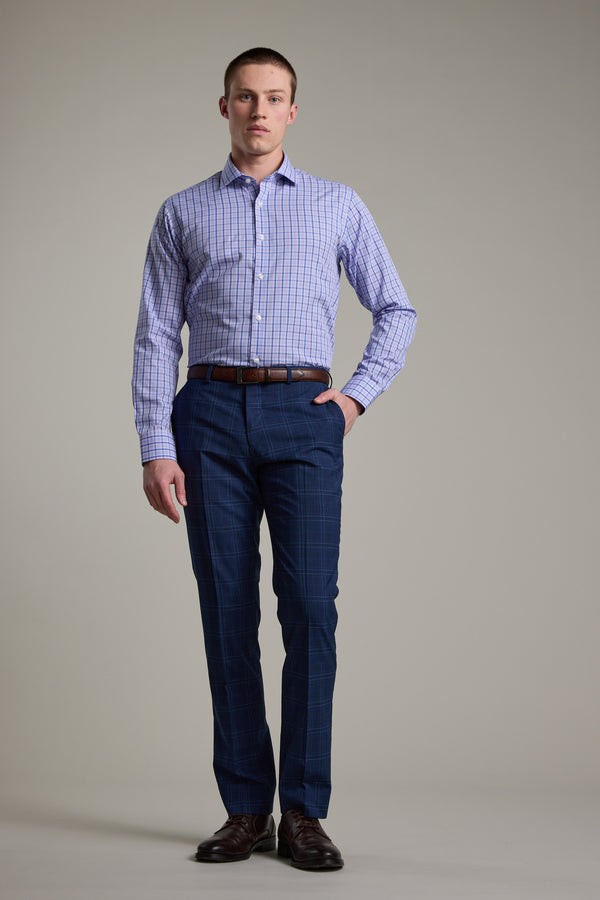 A man stands against a plain background wearing the Barkers Denton Check Business Shirt in light blue, dark blue plaid dress pants, a brown belt, and dark brown dress shoes, with one hand in his pocket.