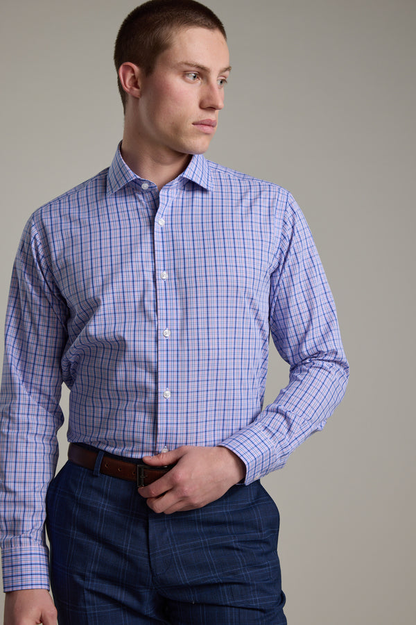 A man in a Barkers Denton Check Business Shirt, tailored fit in light purple and blue, paired with dark blue plaid pants and a brown belt, stands sideways with his left hand on his waist, gazing to the right.