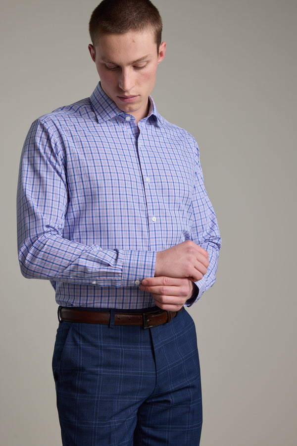 A man wearing the Barkers Denton Check Business Shirt in light purple adjusts his cuff while paired with blue plaid pants, against a plain neutral background.
