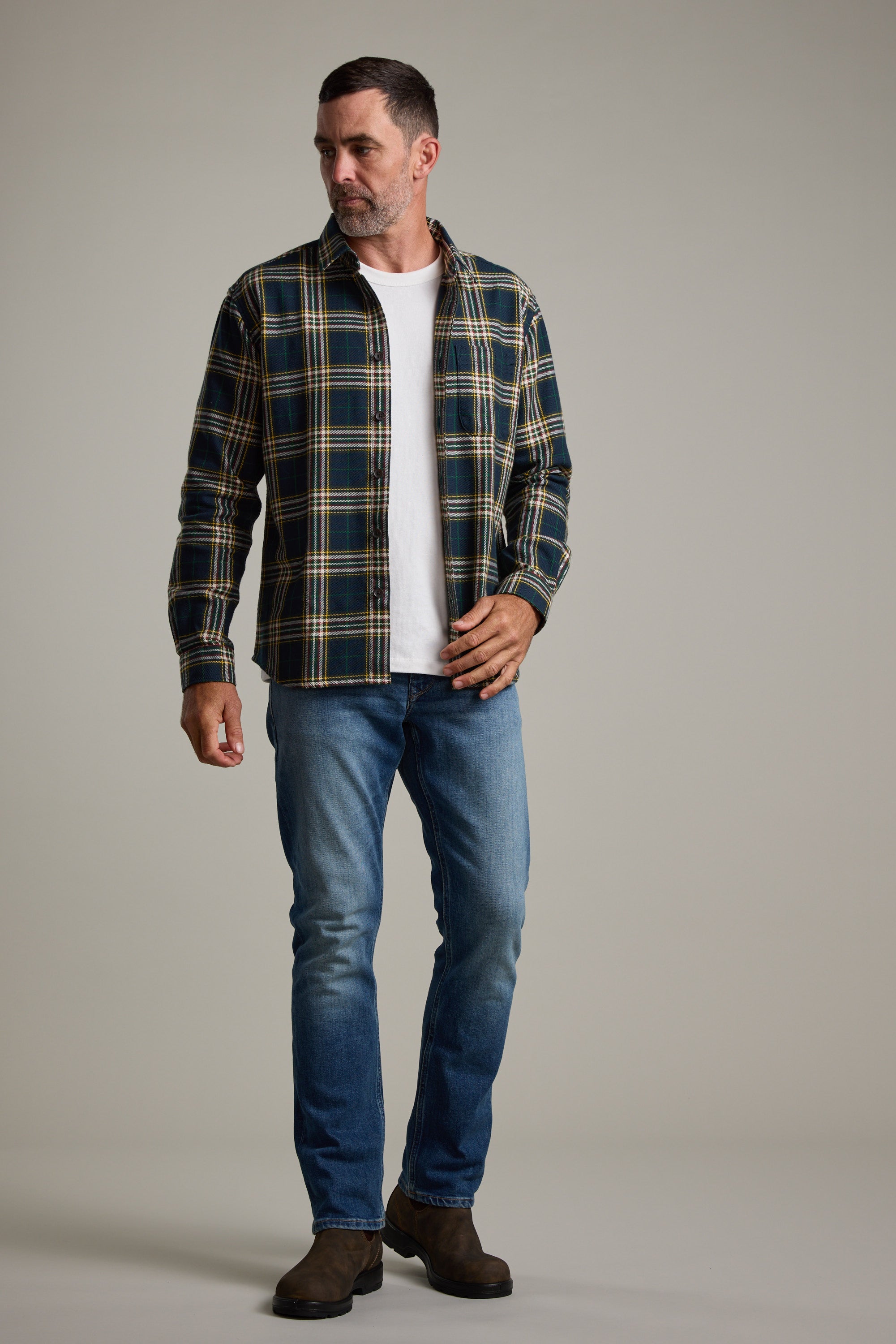 A man stands against a plain light gray background, looking to his left, wearing the Barkers Brennan Check Overshirt in green and yellow plaid over a white t-shirt, blue jeans, and brown boots.