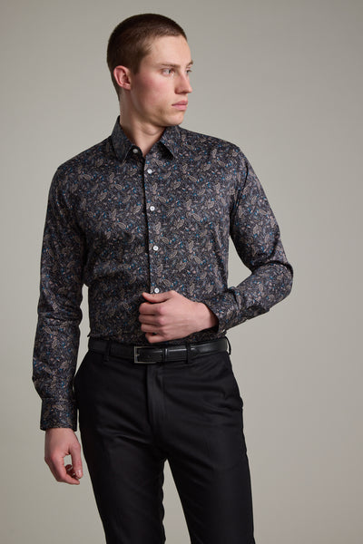 A man in a tailored-fit Barkers Hilton Print Shirt with a subtle floral pattern and black trousers stands against a light gray background, looking left with one hand near his buttons, showcasing a modern, stylish appearance.