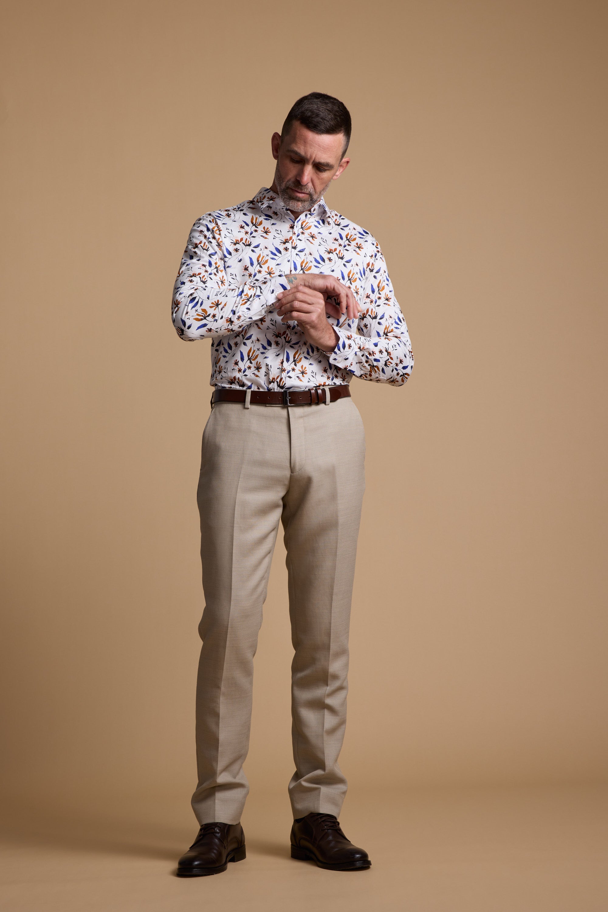 A man wearing the Barkers Milo Print Shirt—a tailored fit with a colorful leaf pattern—paired with beige pants and brown dress shoes, stands against a tan background, looking down as he adjusts his shirt cuff.