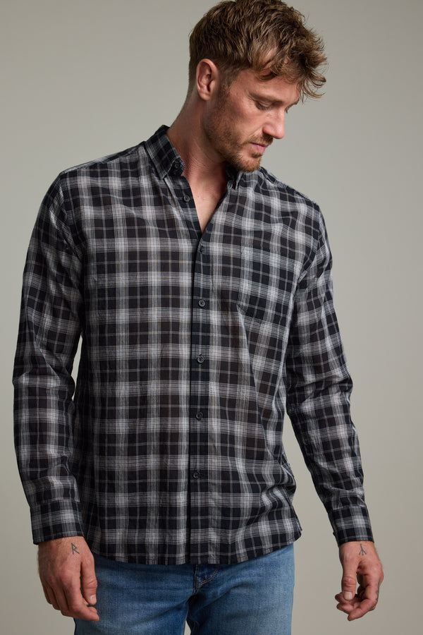 A man with short brown hair and light facial hair wears the Barkers Carter Check Shirt—a classic fit with a black, white, and grey check pattern—paired with blue jeans. He looks downward against a plain background.