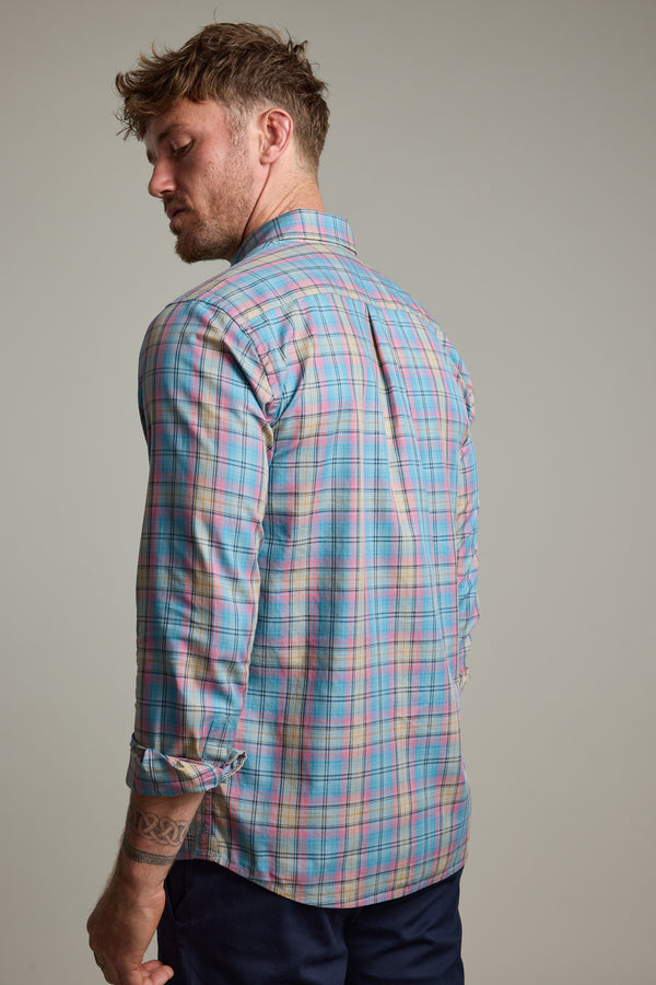 A man with short, tousled hair stands facing away, wearing the Barkers Hayne Check Shirt in pastel plaid with rolled-up sleeves and dark pants. His tattooed left arm hangs loosely by his side.