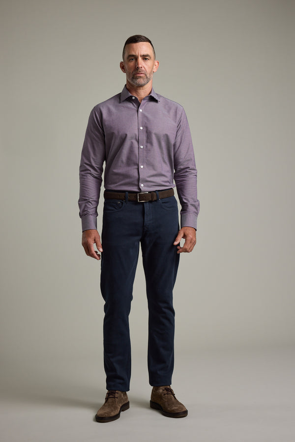 A man with short hair and a beard stands facing forward against a plain gray background, wearing Barkers' Waverly Check Business Shirt in a tailored fit, paired with dark blue pants, a brown belt, and brown suede shoes.