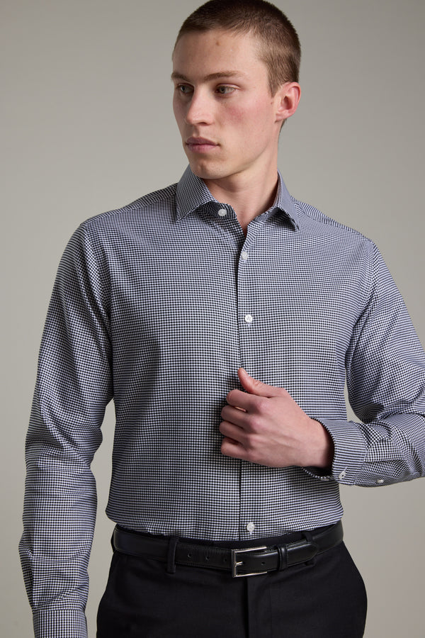 A young man stands against a plain background, looking left with one hand near his chest, wearing the Barkers Mitchel Check Business Shirt and black pants.