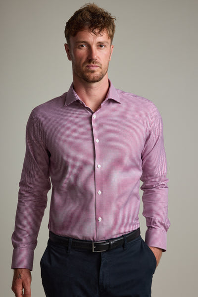 A man with short, wavy brown hair and stubble wears the Barkers Mitchel Check Business Shirt in red and white, made from responsibly sourced cotton. The tailored fit shirt is styled with dark trousers against a plain light background.