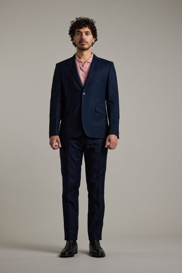 A man with curly hair faces forward, wearing Barkers’ Moris Cotton Pant, a tailored pale pink shirt, and black dress shoes against a light gray background.