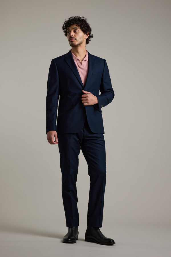 A man with curly hair, dressed in Barkers Moris Cotton Pants, a tailored navy suit, a light pink polo shirt, and black shoes stands against a plain light gray background, gazing slightly to the side.