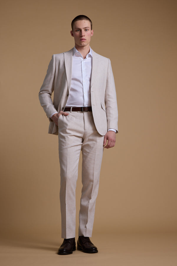 A man stands against a beige background, wearing Barkers’ Somerton Texture Pant in a breathable linen-cotton blend, paired with a white shirt, brown belt, and dark brown shoes. One hand rests in his pocket and he has a neutral expression.