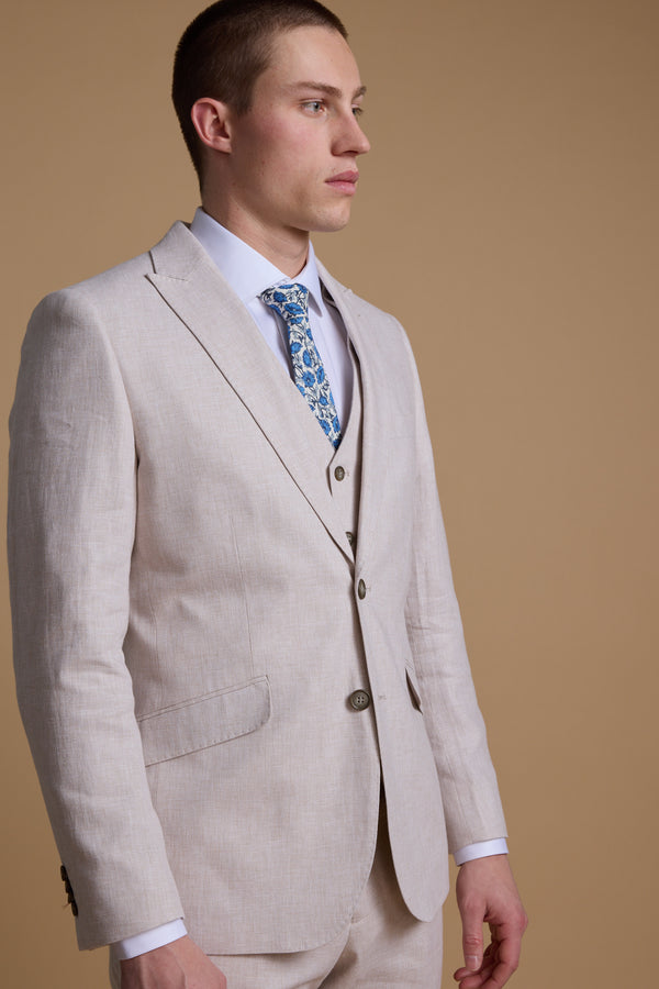 A man in the Barkers Somerton Linen Blazer—tailored fit, light beige—paired with a white shirt and blue floral tie stands against a brown background, looking to the side; ideal summer wedding style.