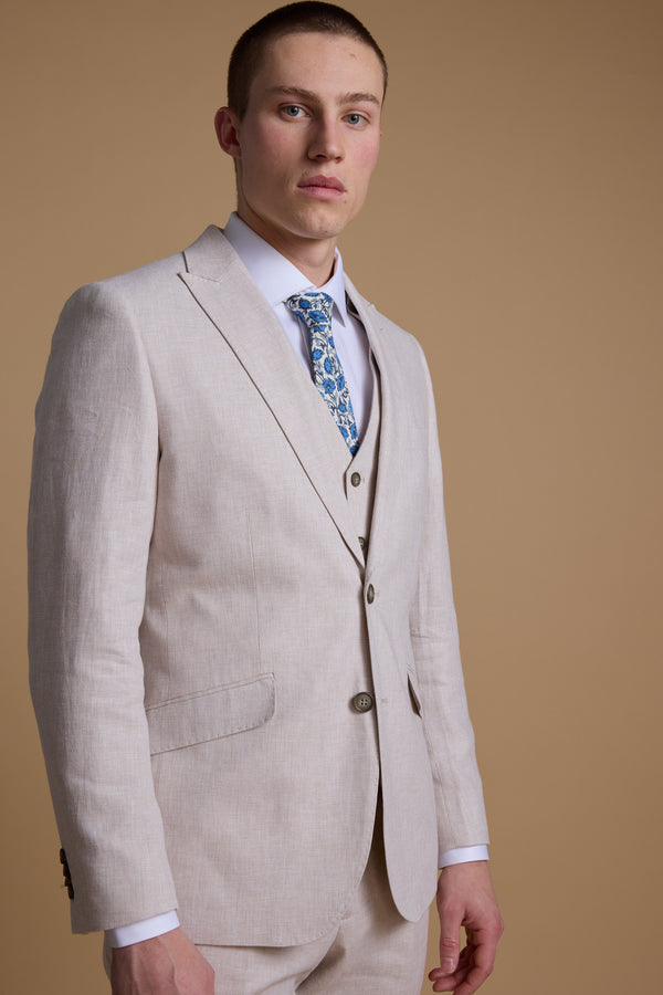 A man in the Barkers Somerton Linen Blazer stands against a tan background, wearing a white shirt and blue patterned tie—his tailored look is ideal for summer weddings.