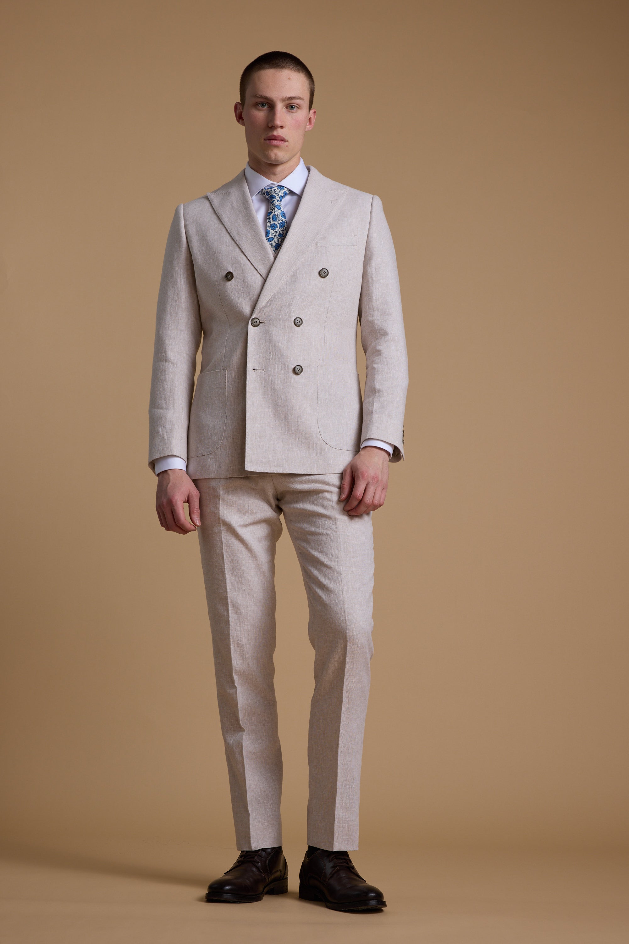 A man wears the Barkers Somerton Linen Double Breasted Blazer with matching trousers, a white shirt, blue patterned tie, and dark brown shoes against a beige background—an elegant summer suit look.