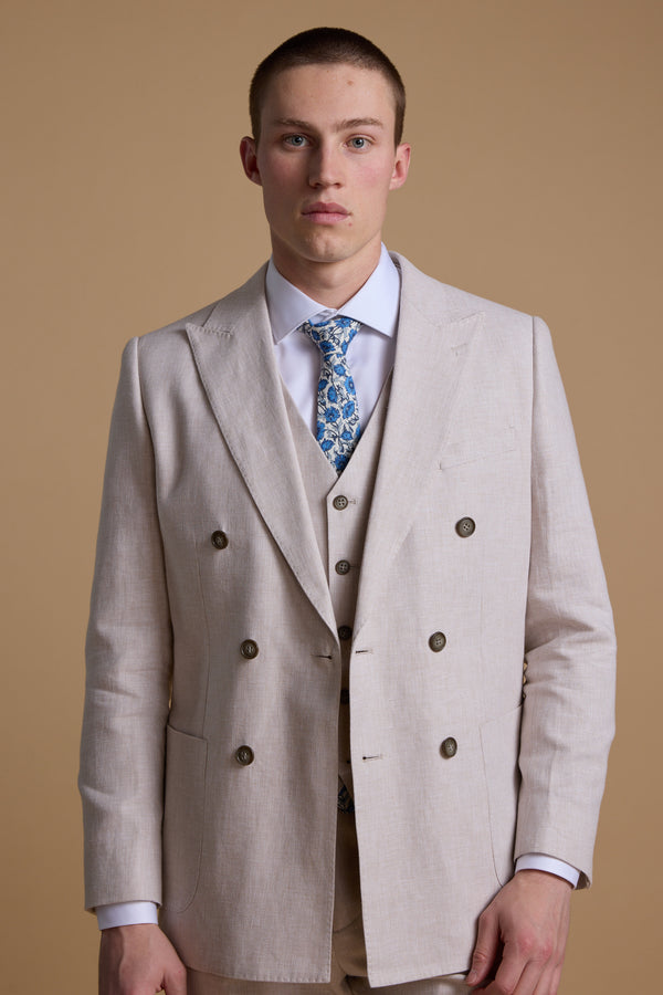 A young man with short hair wears the Barkers Somerton Linen Double Breasted Blazer, paired with a matching vest, white shirt, and blue floral tie for a refined summer look against a plain tan background.