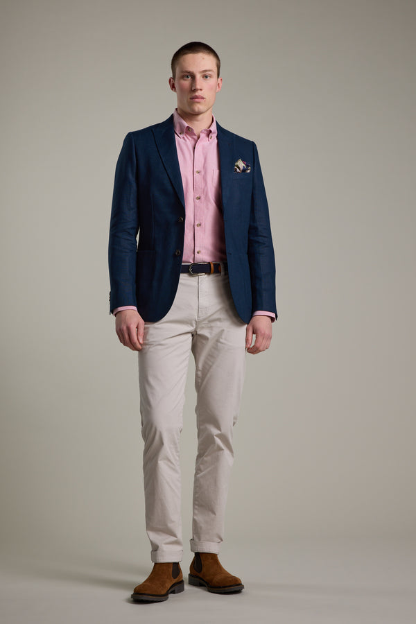 A man stands facing forward in a summer outfit, wearing the Barkers Payson Linen Blazer in navy, paired with a pink shirt, beige pants, brown belt and shoes, and a pocket square. The background is plain and neutral.