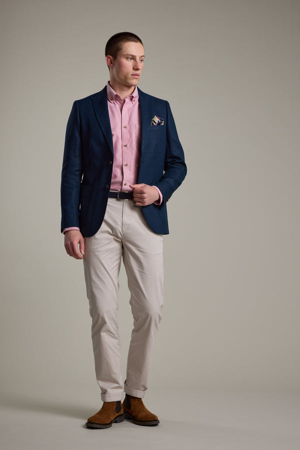 A man stands against a plain background, wearing the Barkers Payson Linen Blazer in navy with a light pink shirt, beige pants, brown shoes, and a patterned pocket square—ideal for effortless summer tailoring—as he looks to the side.