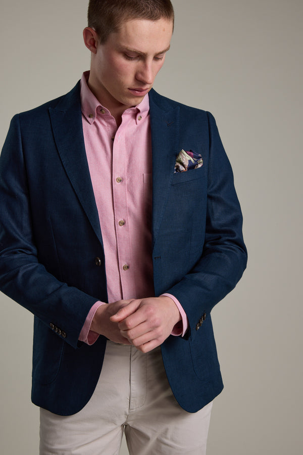 Wearing the Barkers Payson Linen Blazer with a light pink shirt and pale trousers, a man stands against a plain backdrop, looking down with hands clasped. A patterned pocket square completes his refined summer ensemble.