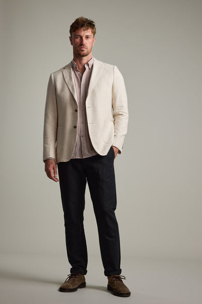A man with short hair and a neutral expression stands against a plain background, wearing the Barkers Bernard Herringbone Blazer over a striped shirt, dark trousers, and brown suede shoes, with one hand in his pocket.