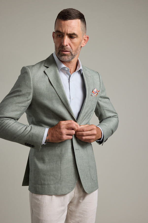 A man with short hair and a beard fastens the Barkers Marion Texture Blazer in green over a light blue dress shirt. He pairs it with light trousers and a patterned pocket square, standing against a neutral background.