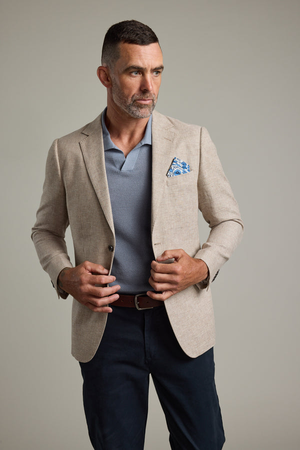 A man with short dark hair and a beard wears the Barkers Welland Texture Blazer in beige over a blue polo and dark pants, accessorized with a blue pocket square, looking to the side against a plain background.