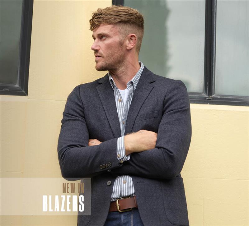 A man with short hair, wearing a dark blazer over a striped shirt and jeans, stands with arms crossed in front of a pale yellow wall and window. Text reads NEW IN BLAZERS.