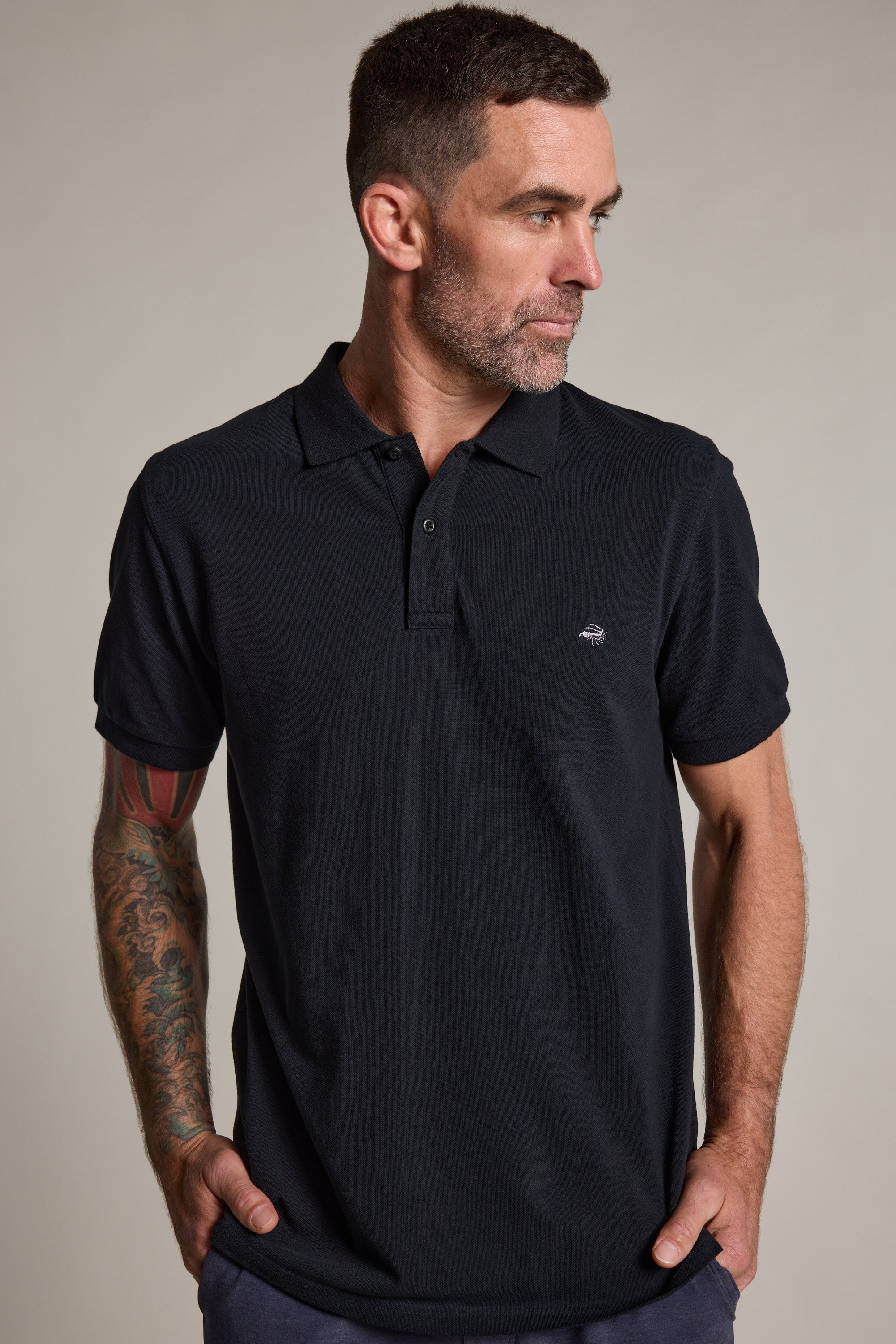 A man with short dark hair, a beard, and a tattoo sleeve on his left arm stands with his hands in his pockets, looking left. He wears a black Barkers Regent Pique Polo featuring a small embroidered logo.