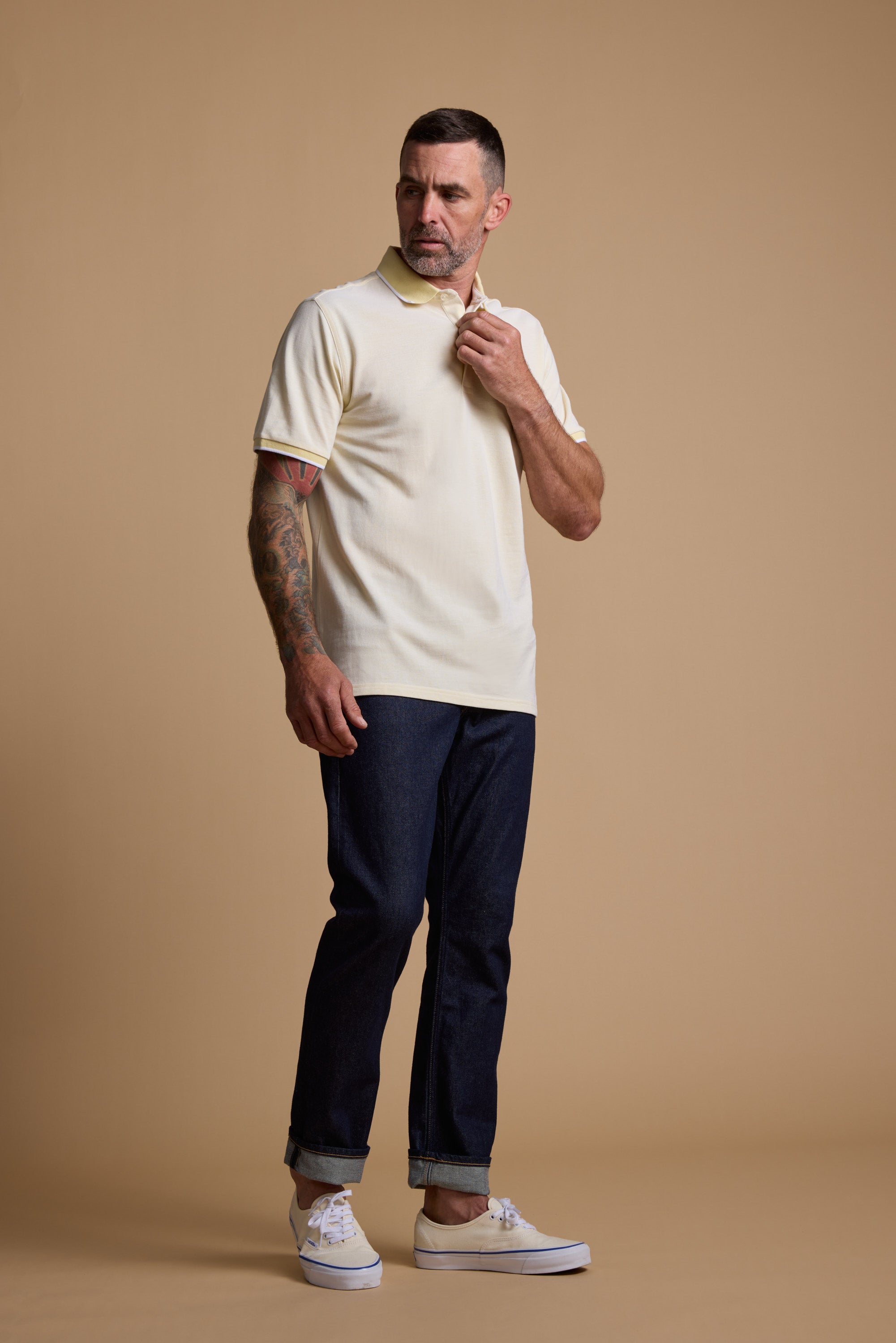 A man with short hair and a tattooed right arm wears the Barkers Birdie Pique Polo in pale yellow, paired with dark cuffed jeans and white sneakers, standing against a plain beige background and looking to his left.