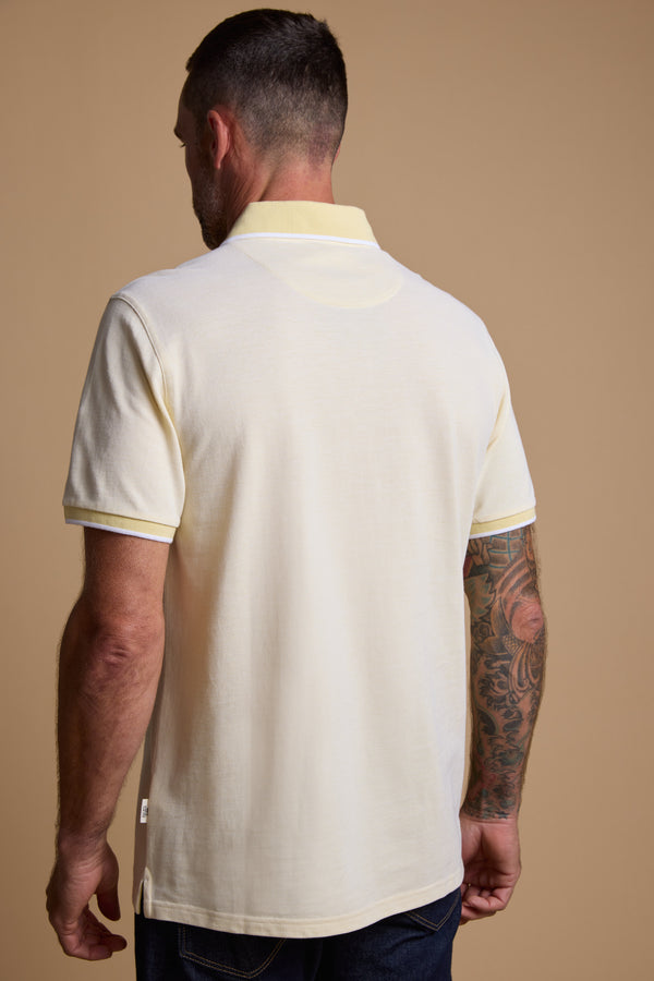 A man with short dark hair and a tattooed right arm is seen from behind, wearing the Barkers Birdie Pique Polo in light yellow and dark jeans, standing against a plain beige background.