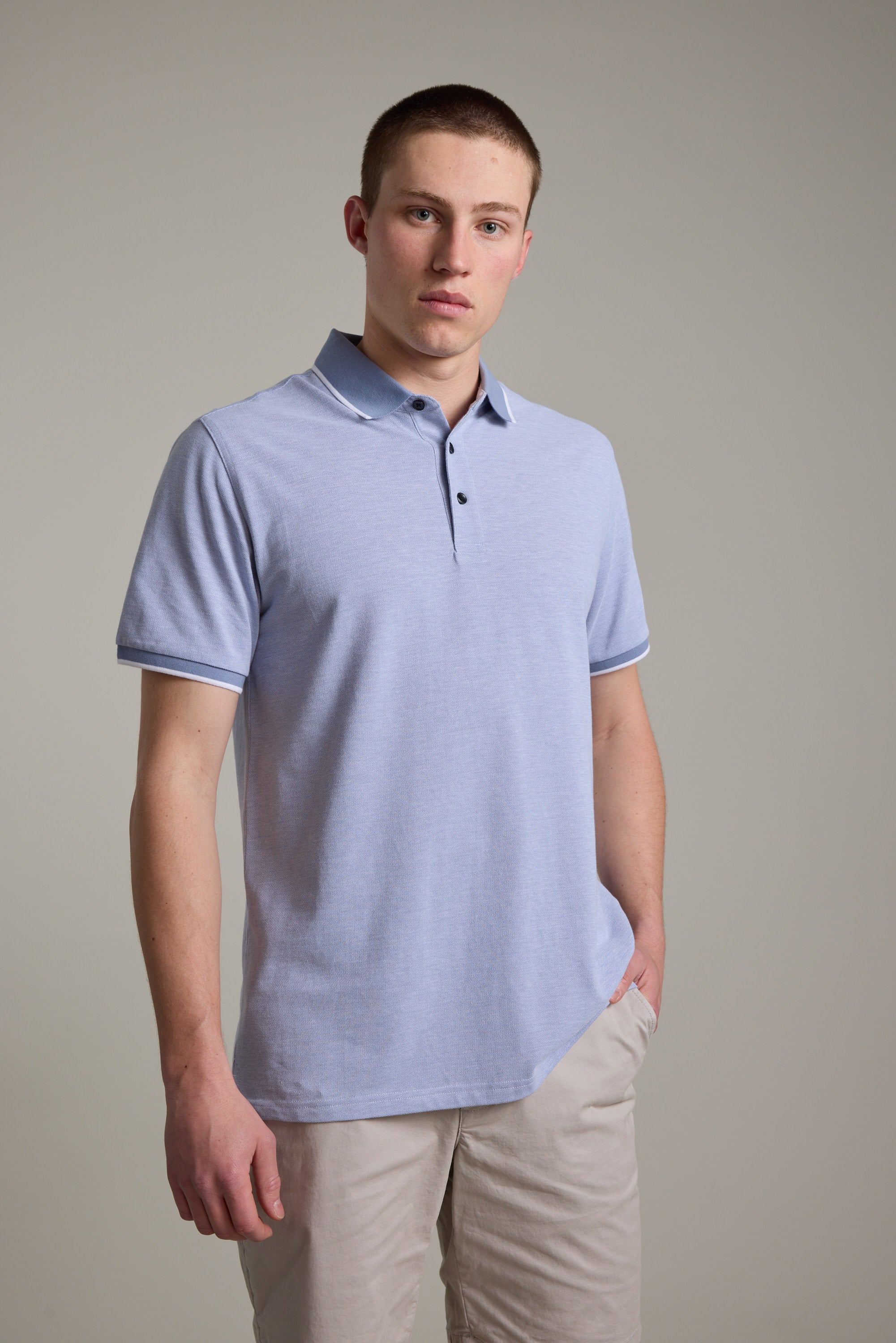 A young man stands against a plain background, wearing the light blue Birdie Pique Polo by Barkers and beige shorts. With short hair and a neutral expression, one hand in his pocket, he showcases this versatile essential for any casual look.