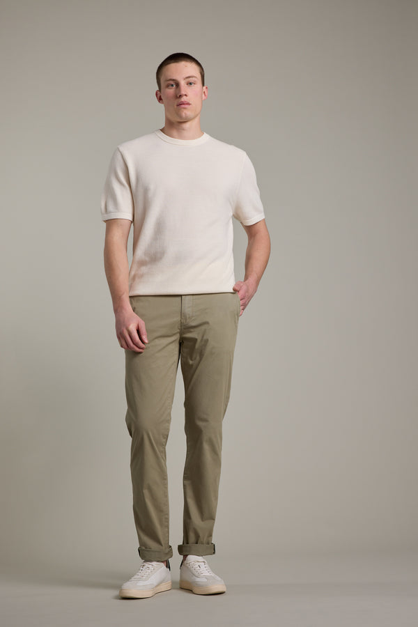 A young man stands facing forward against a plain gray background, wearing Barkers' Flinders Cotton Knit Tee in off-white, paired with light olive-green cuffed pants and white sneakers.
