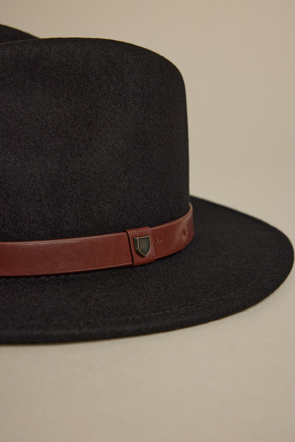 A close-up of the Brixton Messer Fedora Hat—a black wool felt fedora with a wide brim and brown leather band, detailed by a small metallic shield accent. The hat is shown against a plain beige background.