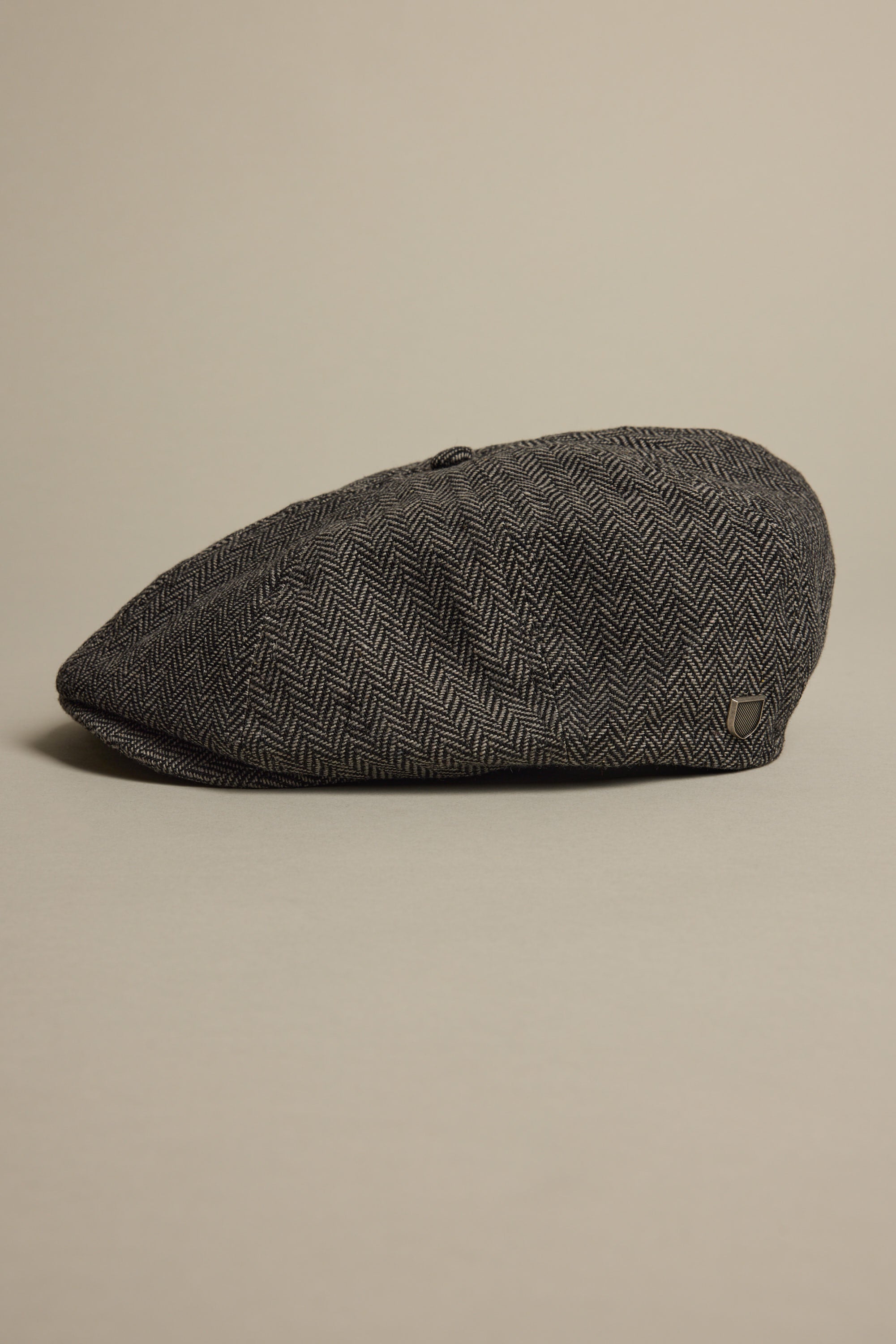 The Brixton Brood Snap Cap by Brixton rests on a light surface, displaying its gray herringbone wool-blend fabric, side profile, and textured pattern.