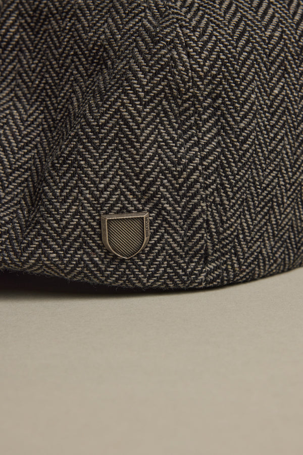 Close-up of the Brixton Brood Snap Cap by Brixton, featuring a textured herringbone wool blend and a small metallic shield emblem near the brim, set against a neutral background.