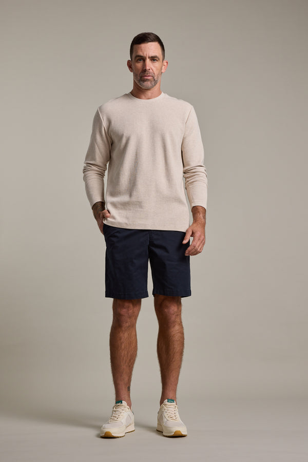 A man stands against a plain background wearing a beige long-sleeve shirt, Barkers Kingly Chino Short in dark navy, and light sneakers. One hand is in his pocket while the other rests at his side.