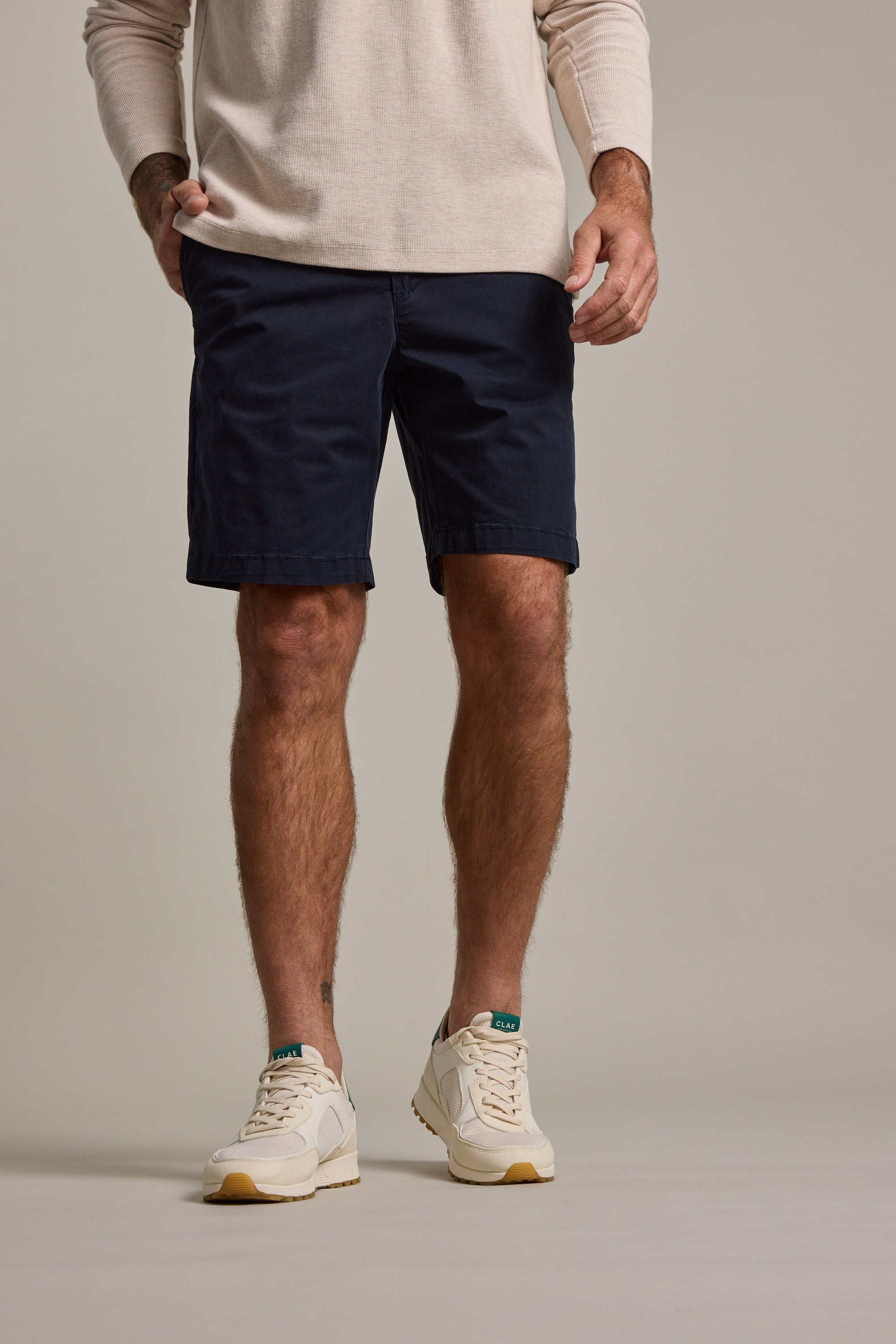 A man, cropped from the shoulders down, stands with hands in pockets, wearing a beige long-sleeve shirt, Barkers Kingly Chino Short men's stretch shorts in navy blue, and white sneakers with green and tan details.