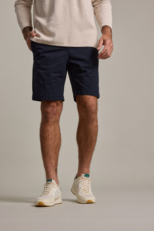 A man, cropped from the shoulders down, stands with hands in pockets, wearing a beige long-sleeve shirt, Barkers Kingly Chino Short men's stretch shorts in navy blue, and white sneakers with green and tan details.