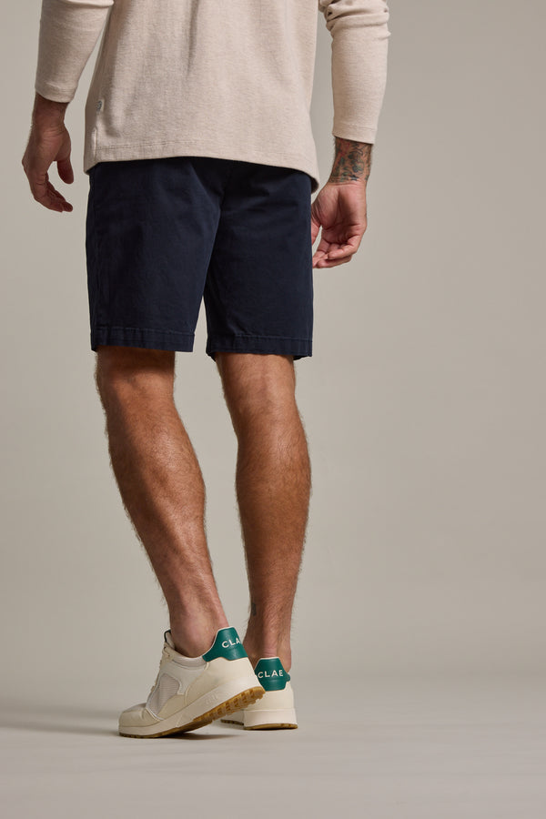 A person is seen from behind wearing a beige long-sleeve shirt, Barkers Kingly Chino Short in navy, and white sneakers with green accents, showcasing the men's stretch shorts against a plain light backdrop.