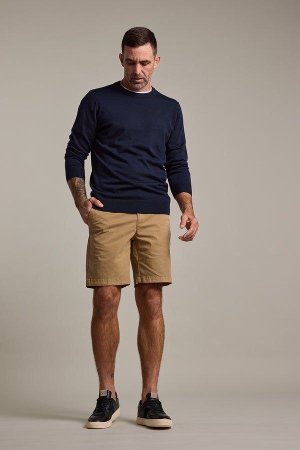 A man with short hair and a mustache stands against a plain background wearing Barkers’ Kingly Chino Short in organic cotton, paired with a navy sweater and black sneakers. One hand is in his pocket, the other rests at his side.