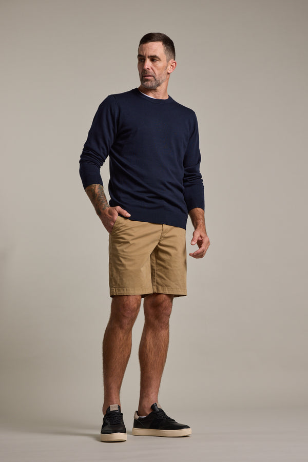 A man stands slightly turned, wearing a navy long-sleeve sweater, black sneakers, and tan Barkers Kingly Chino Shorts for men against a plain light background.