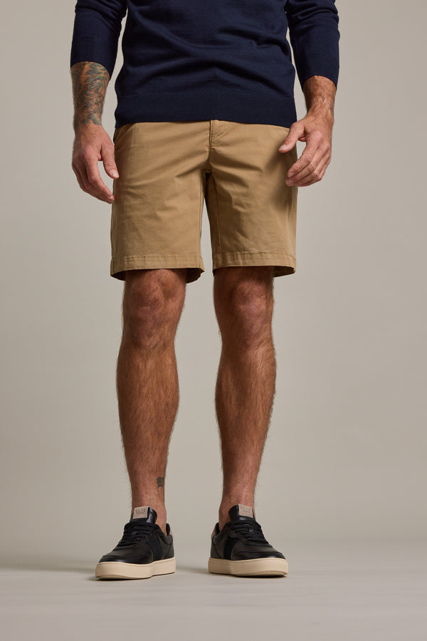 A man wears a navy long-sleeve shirt, black sneakers with white soles, and Barkers Kingly Chino Short—tan men's casual shorts made from organic cotton. His arms are at his sides, showing a tattoo on his left forearm against a neutral background.