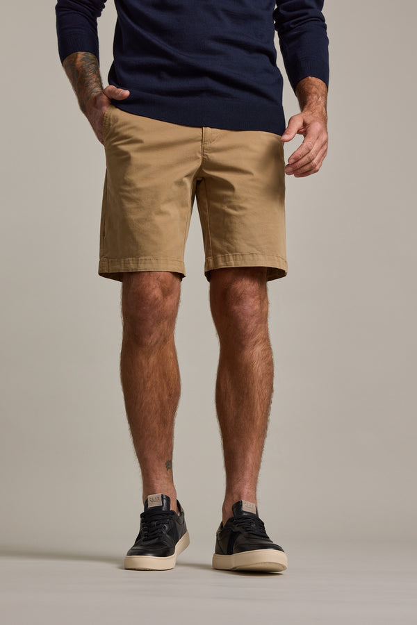 Man wears a navy long-sleeve shirt, Barkers Kingly Chino Short in tan—premium men's shorts made from organic cotton—and black sneakers with white soles, standing against a plain light background. Image shows him from shoulders to feet.
