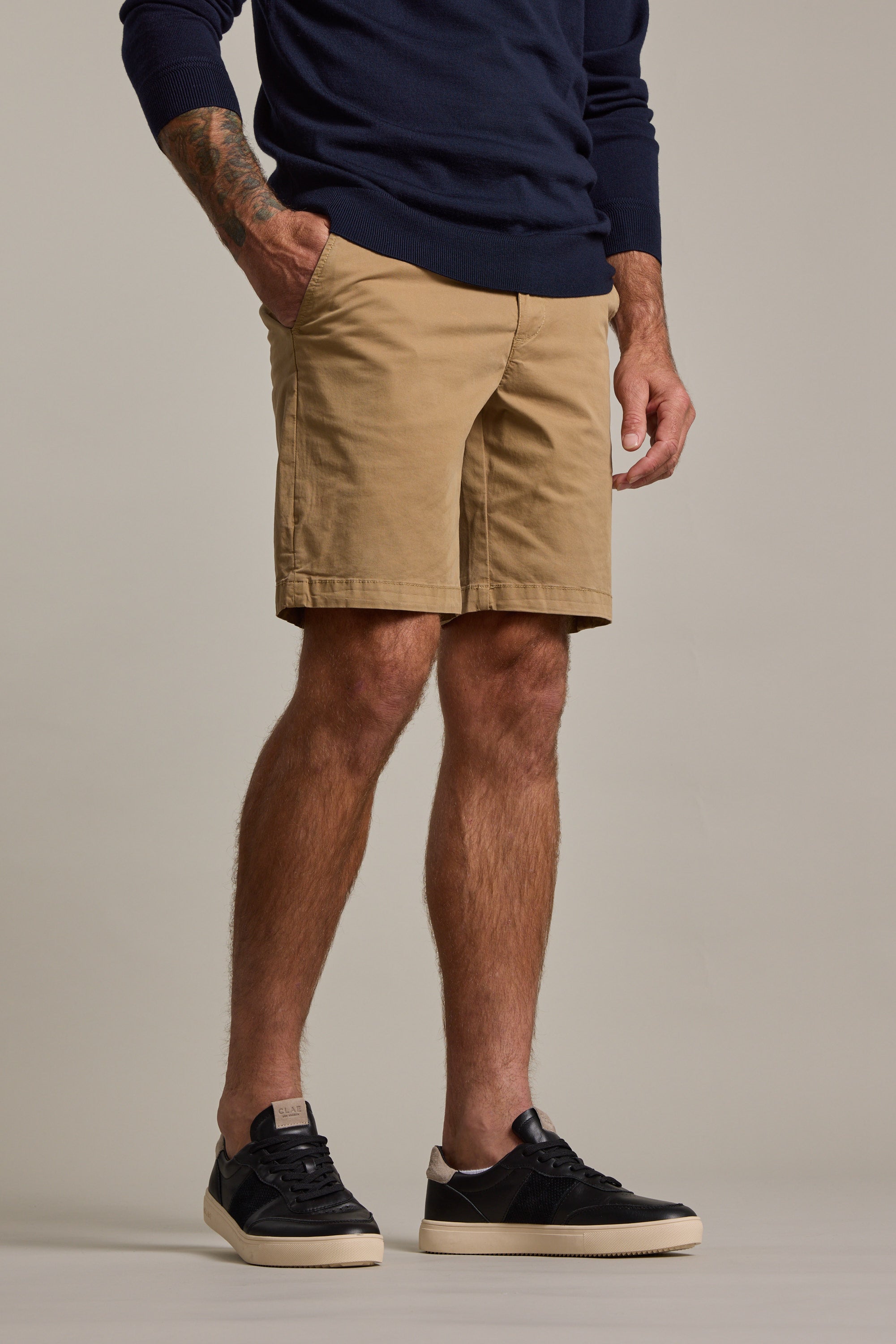 A man wearing a navy sweater, black sneakers, and Barkers Kingly Chino Short men's casual shorts in tan stands with one hand in his pocket against a plain background. His head is not visible in the image.