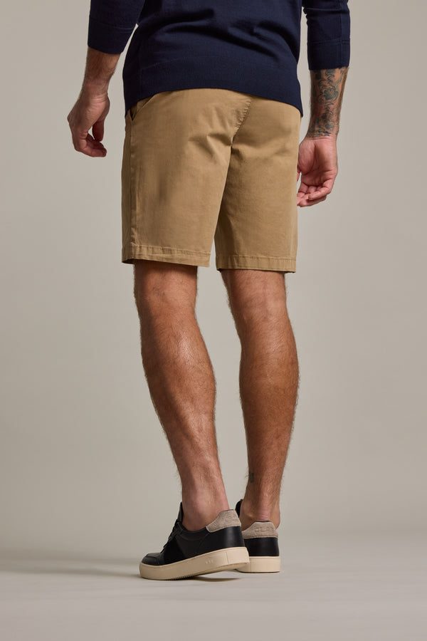 A man with a left forearm tattoo is seen from behind wearing Barkers Kingly Chino Shorts in tan, a navy sweater, and black sneakers with light soles against a plain, neutral background.