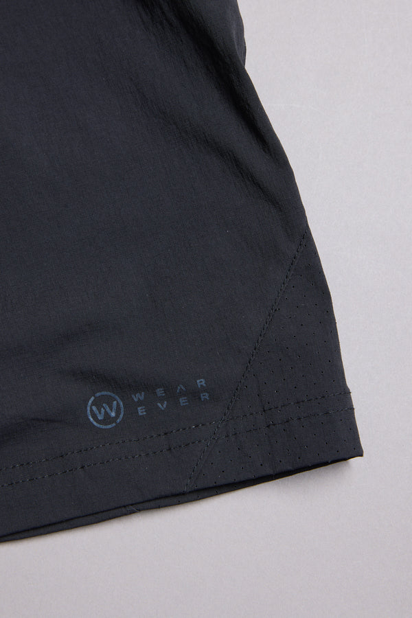 Close-up of the hem on WearEver Agile Short, featuring lightweight stretch fabric, small perforations, and a white WEAREVER logo near the edge, against a plain light gray background.