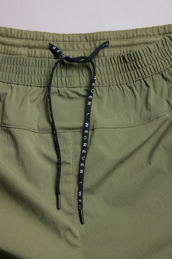 Close-up of WearEver Agile Short, olive green athletic shorts by WearEver with a 7-inch inseam, elastic waistband, and black drawstring bearing white WHEREVER text. The lightweight fabric has a slightly textured look.
