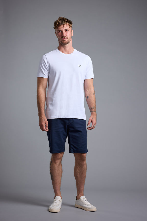 A man wears a white t-shirt, Barkers Anvil 5Pkt Shorts in navy, and white sneakers, standing facing forward against a gray background with a neutral expression and relaxed posture.