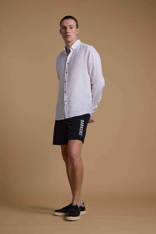 A young man stands against a beige background, wearing a white shirt and Barkers Weekend Shorts by Barkers, made from recycled polyester. He faces slightly to the side with his hands behind his back and is wearing black sneakers.