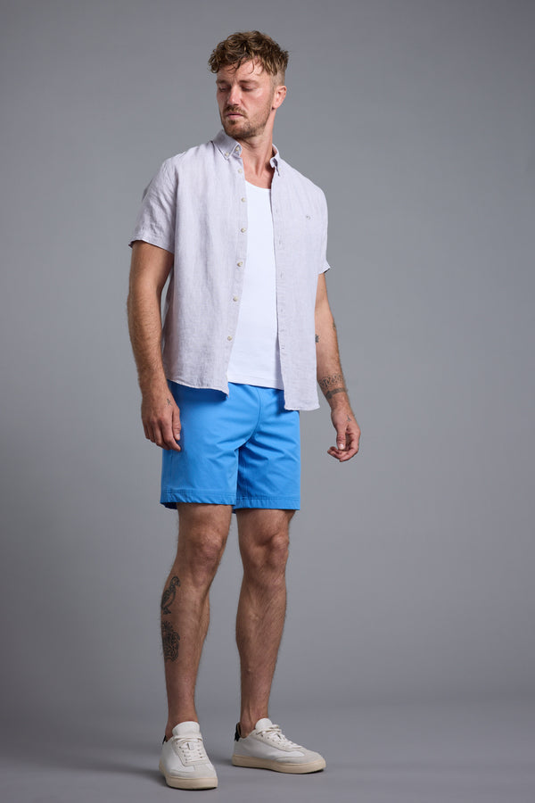 A man stands against a plain gray background wearing a light short-sleeve button-up shirt over a white T-shirt, Barkers Weekend Short in bright blue, and white sneakers. He looks down to his left with a neutral expression.