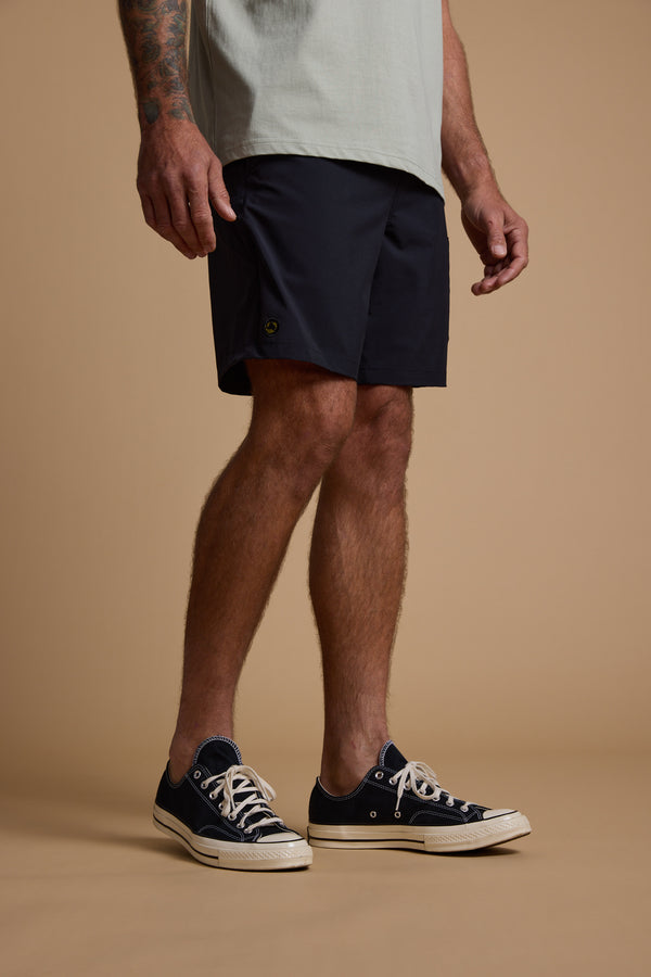 A man stands against a beige background, wearing Legasea Weekend Short and a light green T-shirt with black low-top sneakers. Only his lower body and tattooed left arm are visible.