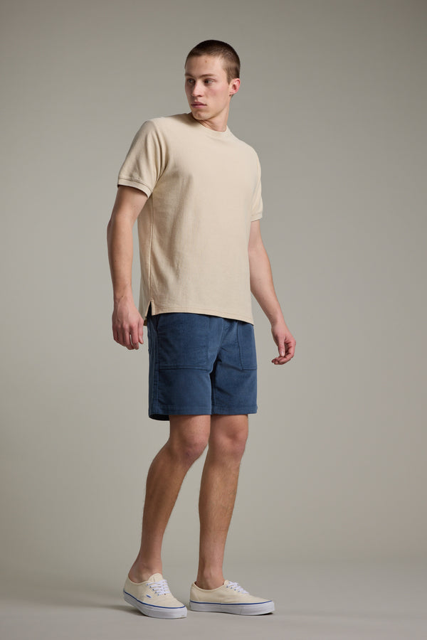 A young man stands against a neutral background, wearing a beige short-sleeve shirt and Barkers Montauk Cord Weekend Short in blue cotton corduroy with an elastic waistband. He looks left, relaxed, in beige canvas sneakers.